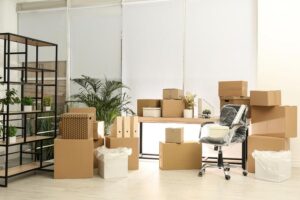 Bureau aménagé avec mobilier de déménagement: cartons empilés, bureau et chaise sous film, étagères et plantes en arrière-plan. Déménagement militaire suggéré par l’arrangement.