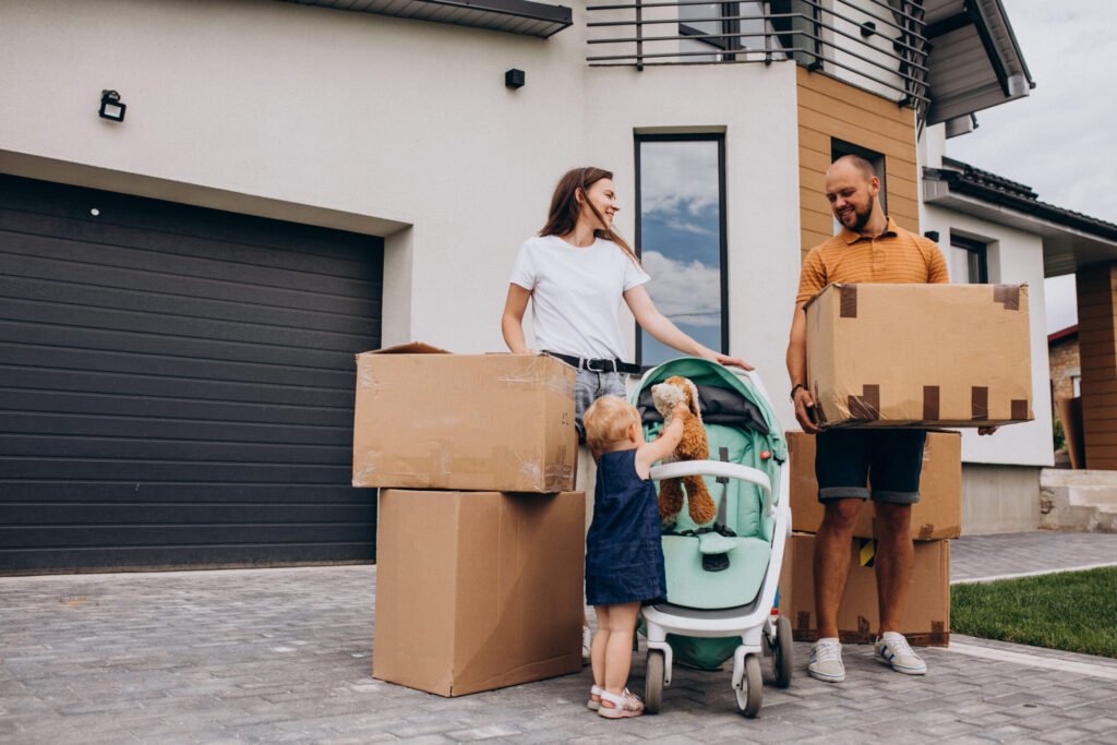 Famille démenageant devant leur maison moderne avec cartons et poussette, prêt à déménager en douceur. Intègre naturellement le guide « comment bien organiser son déménagement en 1 mois ».