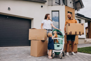 Famille démenageant devant leur maison moderne avec cartons et poussette, prêt à déménager en douceur. Intègre naturellement le guide « comment bien organiser son déménagement en 1 mois ».