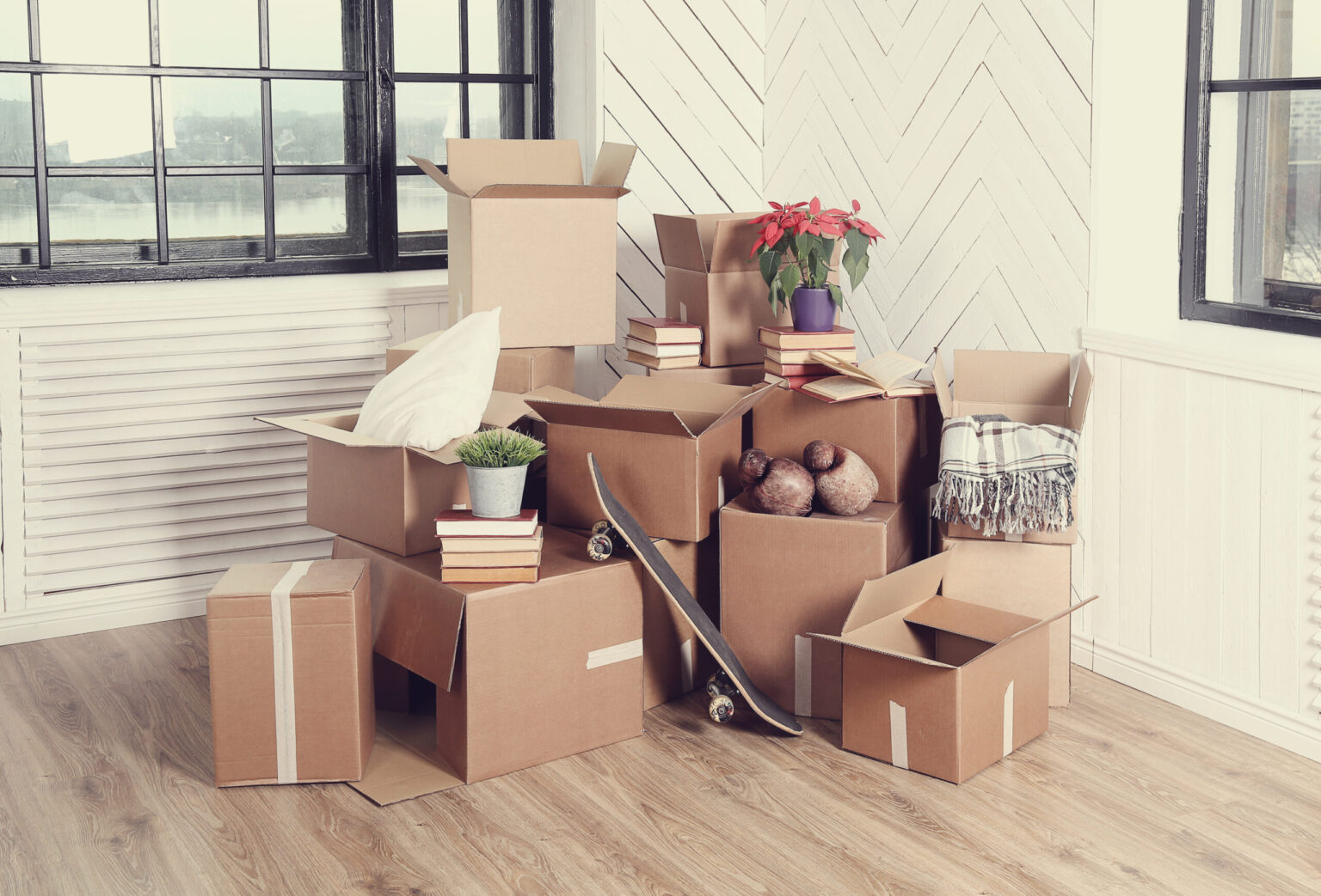 Des cartons en pile dans une pièce lumineuse, avec livres, plantes et une petite planche à roulettes, prêt pour un déménagement. comment préparer son déménagement naturellement inclus.