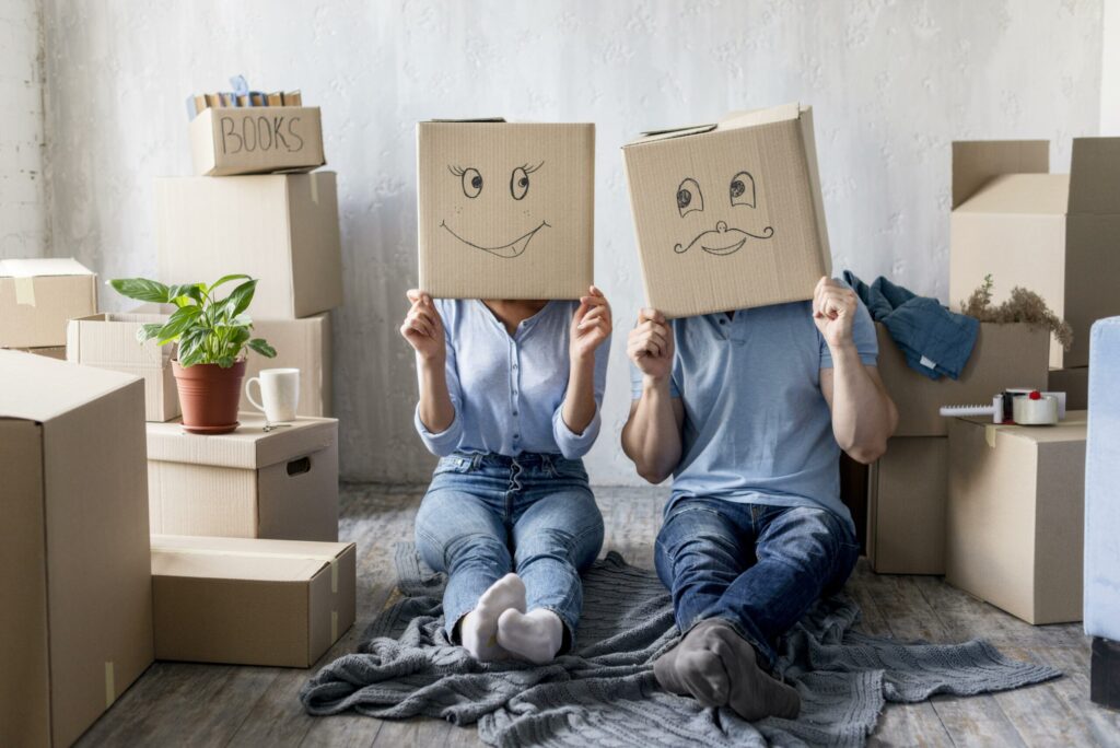 Deux personnes assises sur un tapis, entourées de cartons et de plantes, portent des boîtes sur la tête avec des visages dessinés. Alt text: "checklist déménagement" intégrée? Include keyword naturally. Let's craft: "Deux personnes assises sur un tapis, entourées de cartons et de plantes, portent des boîtes sur la tête dessinant des visages, illustrant une checklist déménagement prête à imprimer." But must be 50-125 chars. Let's count: "Deux personnes assises sur un tapis, entourées de cartons et de plantes, portent des boîtes sur la tête dessinant des visages, illustrant une checklist déménagement." That's long. Let's shorten: "Deux personnes assises sur un tapis, entourées de cartons, portent des boîtes sur la tête avec des visages dessinés, symbole d’une checklist déménagement." Count roughly ~180. Too long. Need 125 max. Try: "Deux personnes assises sur un tapis, entourées de cartons, portent des boîtes sur la tête avec des visages dessinés, symbole d’une checklist déménagement." That's around 140. Hmm. shorter: "Deux personnes assises sur un tapis entourées de cartons portent des boîtes sur la tête avec des visages dessinés, symbole d’une checklist déménagement." Still long. Even shorter: "Deux personnes assises avec des cartons, portent des boîtes sur la tête dessinant des visages, symbole d’une checklist déménagement." This is about 110 chars. Let's deliver. Include keyword exactly "checklist déménagement" integrated: yes as "checklist déménagement" appears. Good. Output only alt text. Let's finalize.