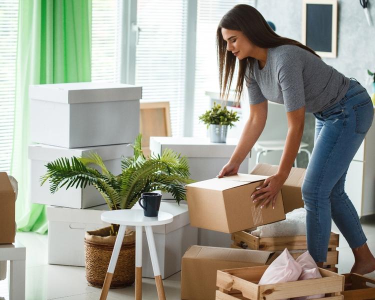 Femme rangeant un carton dans une pièce lumineuse, entourée de boîtes et de plantes, prête à déplacer ses affaires. intégration: comment préparer son déménagement.