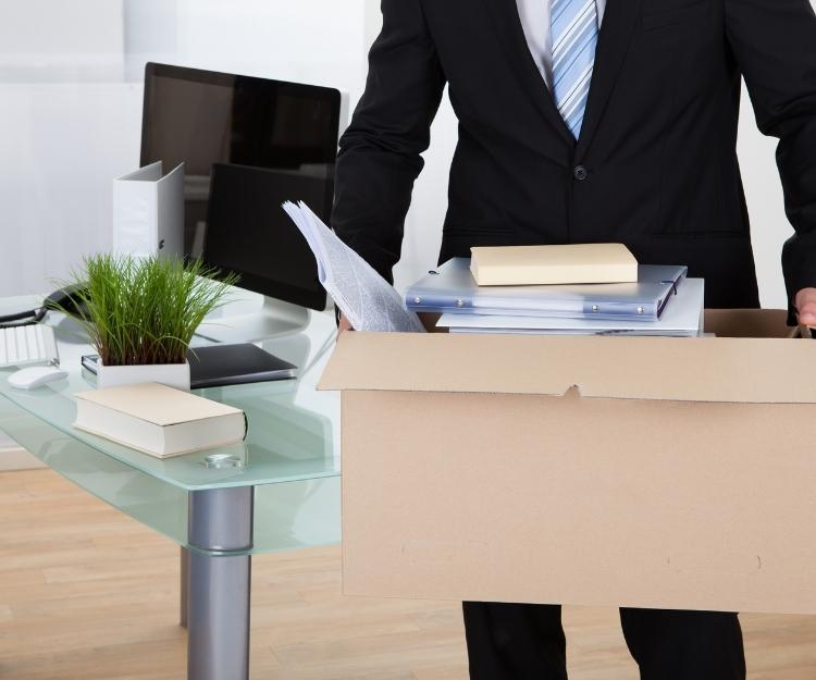 Homme d’affaires en costume avec une boîte contenant des documents et des dossiers près d’un bureau, prêts pour un déménagement, relié à une checklist déménagement.